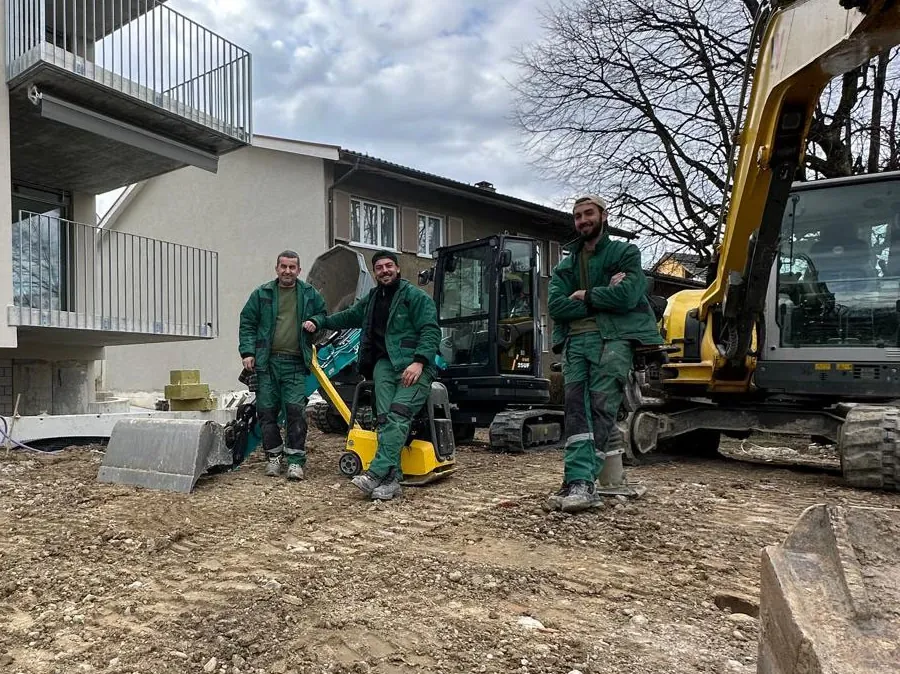 Drei Mitarbeiter in grüner Arbeitskleidung mit Bagger und Rüttelplatte auf einer Baustelle